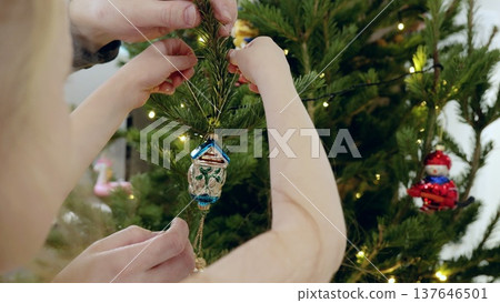 Father and daughter hanging delicate glass house ornament on pine branches, sharing festive christmas tree decorating moment with soft seasonal warmth 137646501
