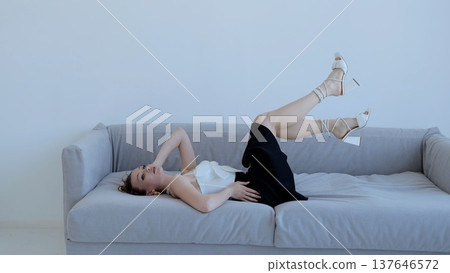 Fashionable model reclining on gray sofa, wearing white top, black skirt, white high heels, legs elegantly raised in sophisticated pose Fashionable model reclining on gray sofa, wearing white top, black skirt, white high heels, legs elegantly raised in sophisticated pose 137646572