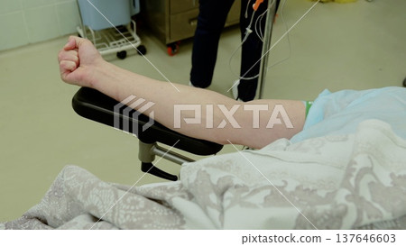 Medical staff preparing patient with clenched fist for surgery, iv drip nearby, hospital operating room setting with medical equipment in background Medical staff preparing patient with clenched fist for surgery, iv drip nearby, hospital operating room setting with medical equipment in background 137646603
