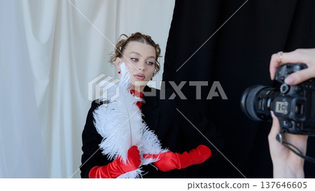 Fashion model posing with white feather accessory, draped in elegant red gloves against black and white background during professional photoshoot Fashion model posing with white feather accessory, draped in elegant red gloves against black and white background during professional photoshoot 137646605