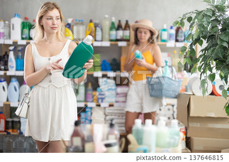 Young girl chooses detergent at home goods store, while in background shoppers browse products Young girl chooses detergent at home goods store, while in background shoppers browse products 137646815