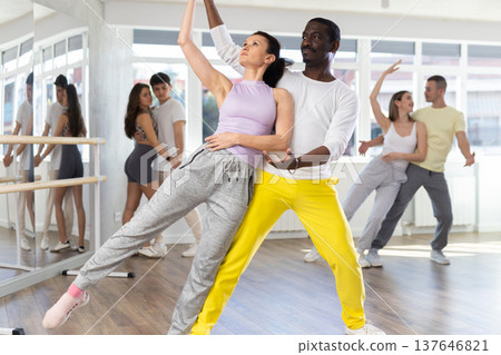 African american man with european girl and group of young active sports people practicing rock and roll dance in dance hall African american man with european girl and group of young active sports people practicing rock and roll dance in dance hall 137646821