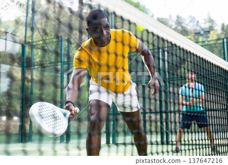 Athletic african american man plays padel. View through the tennis net Athletic african american man plays padel. View through the tennis net 137647216