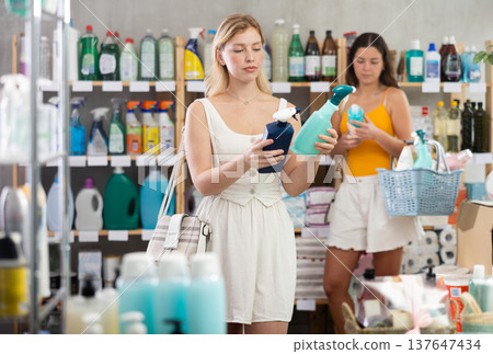 Female shopper chooses spray glass cleaner for windows at home goods store, while in background shoppers browse products Female shopper chooses spray glass cleaner for windows at home goods store, while in background shoppers browse products 137647434
