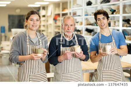 Three positive potters stand in pottery workshop and show created work, finished decorative jug 137647435