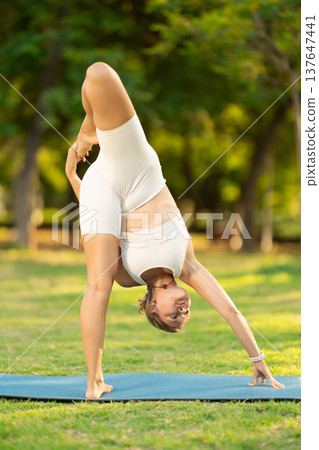 Girl in park on green grass of lawn performs Utthita Hasta Padangushthasana Girl in park on green grass of lawn performs Utthita Hasta Padangushthasana 137647441