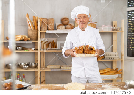 Smiling elderly baker in uniform stands behind counter in small private bakery offering croissants Smiling elderly baker in uniform stands behind counter in small private bakery offering croissants 137647580
