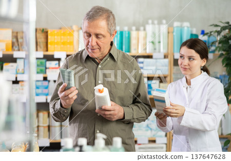 Senior man buys cream at drug store, girl pharmacist in background Senior man buys cream at drug store, girl pharmacist in background 137647628