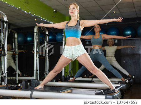 Young woman standing on pilates reformer, perform exercises Young woman standing on pilates reformer, perform exercises 137647652