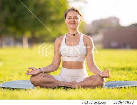 Girl in park on green grass of lawn performs lotus position, padmasana Girl in park on green grass of lawn performs lotus position, padmasana 137647659