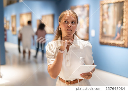 Young girl holding an information booklet looks at paintings in an art exhibition in museum Young girl holding an information booklet looks at paintings in an art exhibition in museum 137648084