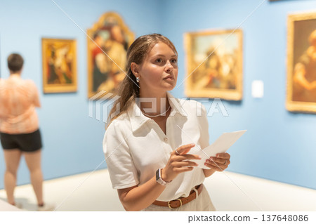 Young girl holding an information booklet looks at paintings in an art exhibition in museum Young girl holding an information booklet looks at paintings in an art exhibition in museum 137648086