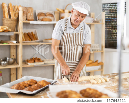 Senior man works in bakery as baker, cuts dough into portions, forms pieces of dough to create buns. Senior man works in bakery as baker, cuts dough into portions, forms pieces of dough to create buns. 137648211