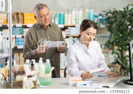 Woman employee at counter with computer sells pills and ointments to senior male customer 137648215