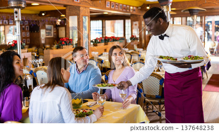 African american waiter serving dishes to cheerful group of adults in restaurant 137648238