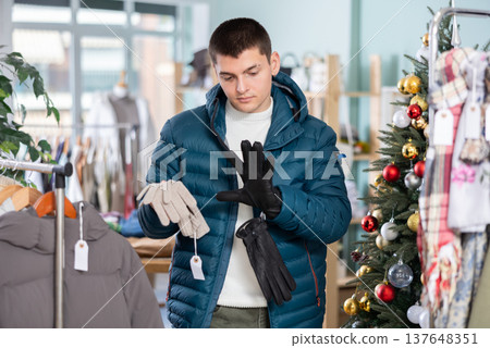 Guy is standing in a store against the background of a Christmas tree and looking at gloves Guy is standing in a store against the background of a Christmas tree and looking at gloves 137648351