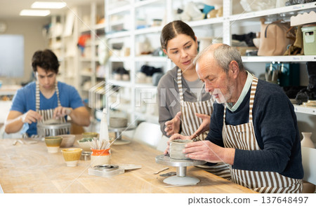 Young woman helping eldery man sculpting ceramic piece Young woman helping eldery man sculpting ceramic piece 137648497