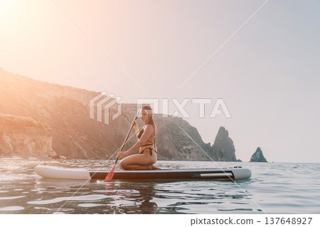 Woman Paddleboarding Ocean Coastline - Woman sits on a paddleboard in the ocean with a rocky coastline in the background. 137648927