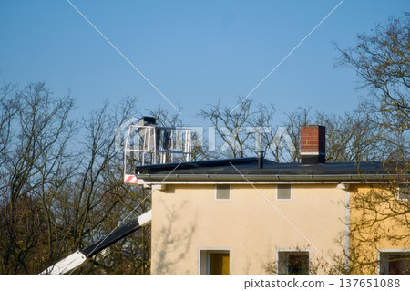 A professional chimney sweep working from an aerial lift platform to inspect and maintain a brick chimney on a residential house roof. A clear view of the specialist at work, emphasizing safety A professional chimney sweep working from an aerial lift platform to inspect and maintain a brick chimney on a residential house roof. A clear view of the specialist at work, emphasizing safety 137651088