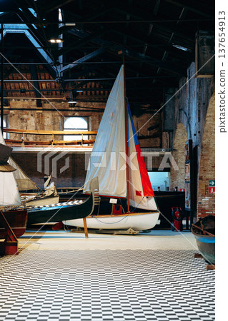 Sailboats and wooden boats inside a large boat house with checkered floor Sailboats and wooden boats inside a large boat house with checkered floor 137654913