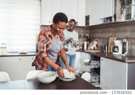 Two cheerful women putting away clean dishes after unloading dishwasher in modern kitchen 137656502