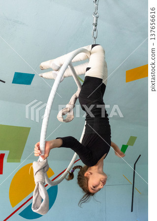 School-age girl in black sportswear doing aerial stunts with hoop, acrobatic pose, elastic bandages for safety, circus artistry. School-age girl in black sportswear doing aerial stunts with hoop, acrobatic pose, elastic bandages for safety, circus artistry. 137656616