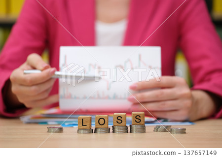 Businesswoman analyzes financial data while addressing losses with wooden blocks on a table filled with documents and coins Businesswoman analyzes financial data while addressing losses with wooden blocks on a table filled with documents and coins 137657149