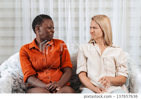 Two women looking at each other with disgust, sitting on a sofa Two women looking at each other with disgust, sitting on a sofa 137657636