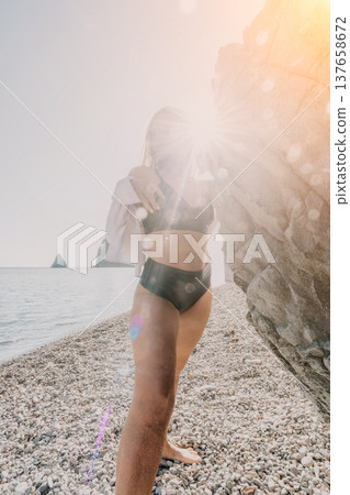 Woman beach bikini standing on a scenic rocky shoreline with bright sun flare and ocean waves during summer vacation Woman beach bikini standing on a scenic rocky shoreline with bright sun flare and ocean waves during summer vacation 137658672