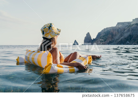 Woman, Beach, Relaxation: A woman in a hat floats on an inflatable raft in the ocean, enjoying the view of a rocky coastline. Woman, Beach, Relaxation: A woman in a hat floats on an inflatable raft in the ocean, enjoying the view of a rocky coastline. 137658675