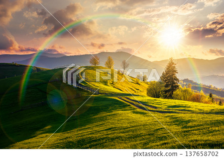 sunset over countryside landscape in mountains. rural picture of wooden fence near dirt road through grassy fields on rolling hills. scenery with distant ridge during springtime weather with rainbow 137658702