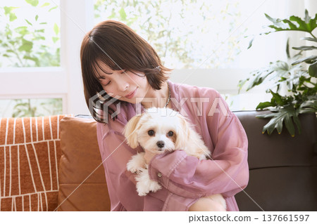 一位女士抱著一隻馬爾濟斯犬；與寵物共度輕鬆時光；在客廳裡與狗狗一起生活。 137661597