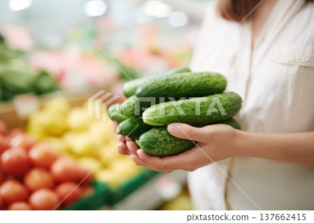 Food shopping. Focus on hands holding clean green cucumbers in supermarket grocery store aisle. Blurred fruits and vegetables shelves background. Fresh Produce Section. Woman choosing buying groceries Food shopping. Focus on hands holding clean green cucumbers in supermarket grocery store aisle. Blurred fruits and vegetables shelves background. Fresh Produce Section. Woman choosing buying groceries 137662415