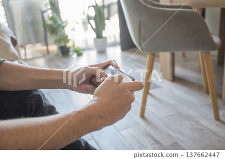 A teenager sits on a couch in a living room playing a video game on a mobile phone. A teenager sits on a couch in a living room playing a video game on a mobile phone. 137662447