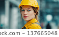 A young Caucasian woman with brown hair wears a yellow hard hat and a work jacket. She stands confidently in a construction environment, symbolizing strength and equality. 137665048