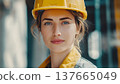 A young Caucasian woman with long brown hair wears a yellow hard hat and a work jacket. She stands confidently at a construction site, symbolizing strength and equality. 137665049