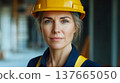 A confident woman wearing a yellow hard hat and blue work uniform stands in a construction site. She represents strength and equality in atypical professions. 137665050