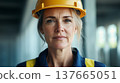A confident middle-aged Caucasian woman wearing a yellow hard hat and blue work uniform stands in a construction site. She represents strength in atypical female professions. 137665051