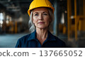 A senior Caucasian woman with gray hair wears a yellow hard hat and blue work uniform. She stands confidently in a construction site, symbolizing strength and equality. 137665052