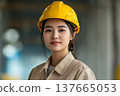 A young Asian woman wearing a yellow hard hat and beige work uniform stands confidently in a construction site. She represents strength in atypical female professions. 137665053