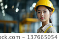 A young Asian woman wearing a yellow hard hat and work uniform stands confidently in a construction site, symbolizing strength and equality in atypical professions. 137665054