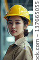 A young Asian woman wearing a yellow hard hat and beige work uniform stands confidently in a construction environment, symbolizing strength and equality in atypical professions. 137665055