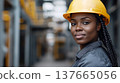 A confident young African woman with braided hair wears a yellow hard hat and gray work uniform. She stands in a construction environment, symbolizing strength and equality. 137665056