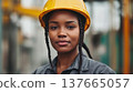 A young African woman wearing a yellow hard hat and gray work uniform stands confidently at a construction site. She represents strength in atypical female professions. 137665057
