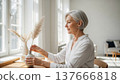 Elegant mature European woman arranging pampas grass in ceramic vase by bright kitchen window wearing white linen shirt 137666818