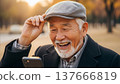Happy Asian elderly man with white beard wearing flat cap laughing while using smartphone outdoors in golden sunlight 137666819