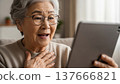 Happy elderly Asian grandmother using tablet device for video call at home wearing glasses and beige sweater 137666821