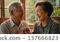 Happy senior asian couple holding hands laughing together by sunny window in cozy home interior with warm natural lighting 137666823