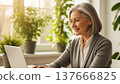 Mature European woman with grey hair using laptop at home office desk surrounded by green plants near bright window 137666825