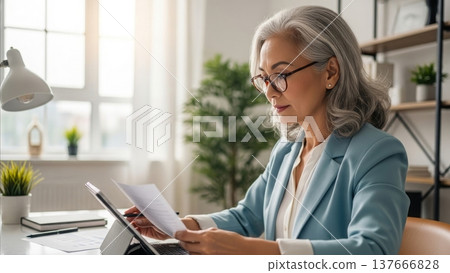Professional mature businesswoman with silver gray hair reading documents at office desk with laptop computer and plants 137666828